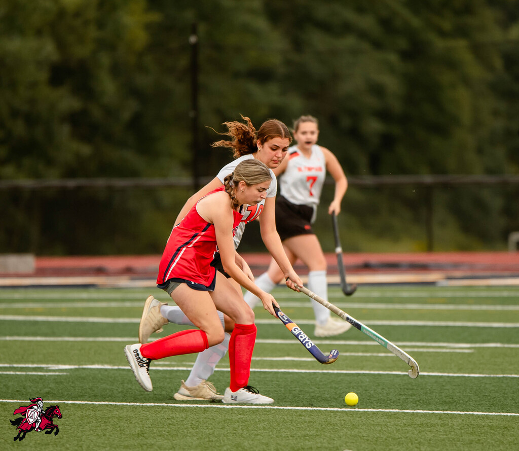 Three players in sports uniforms on a field, two playing field hockey with sticks, one with a ball.