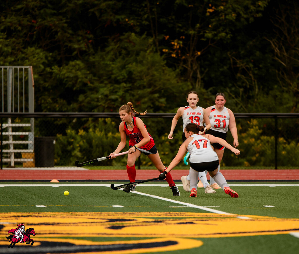 A group of four women play field hockey outdoors. One woman has a ball, and the others are close.