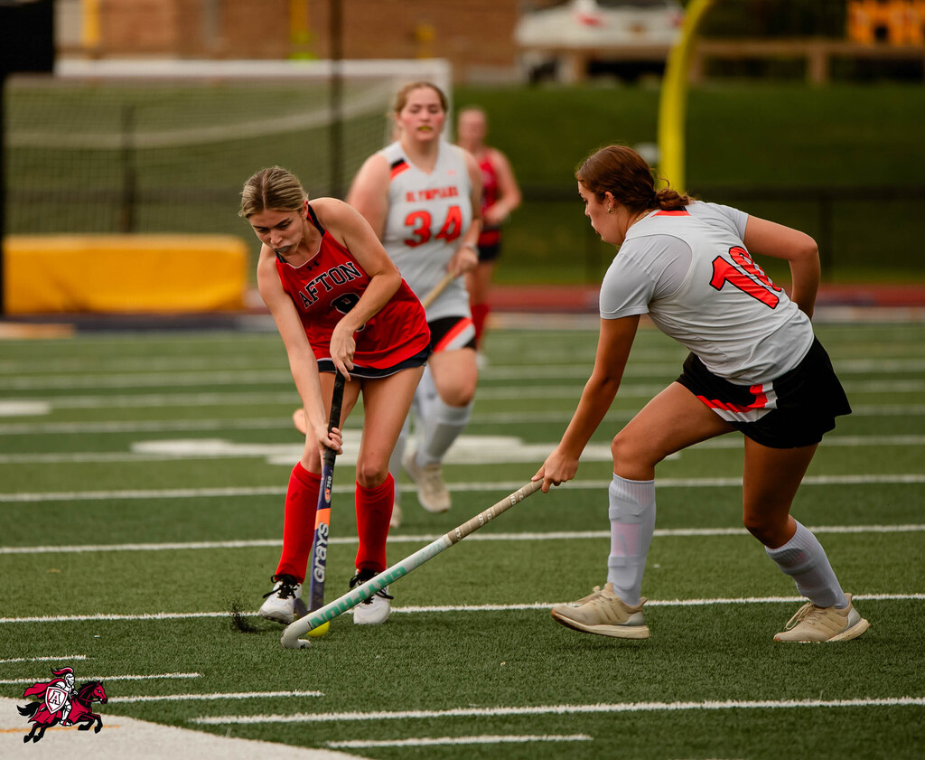 Four women play field hockey on a field. One holds a hockey stick, another extends one.