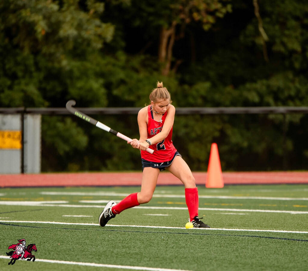 A woman swings a hockey stick on a sports field, wearing red athletic gear. An orange cone is behind her.