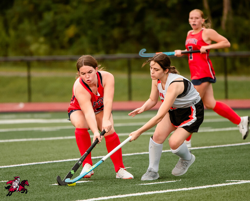 Three women play field hockey on a field. One woman in red is poised to strike.