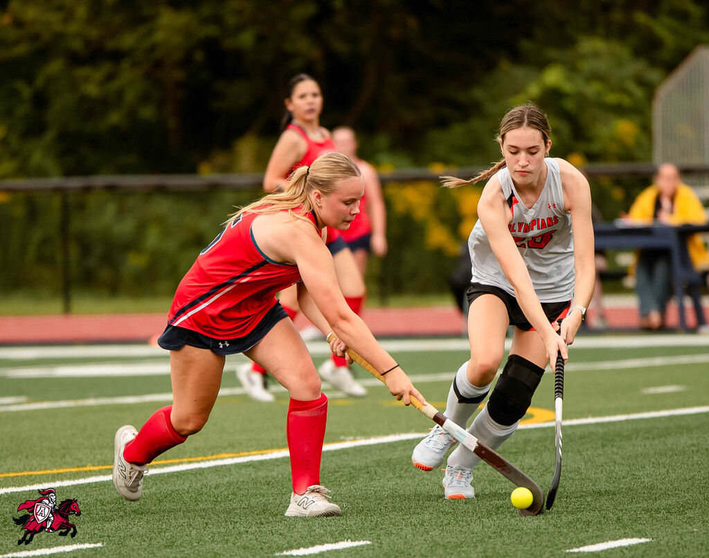 Two women playing field hockey on a grassy field with white lines. One in red, the other in gray.
