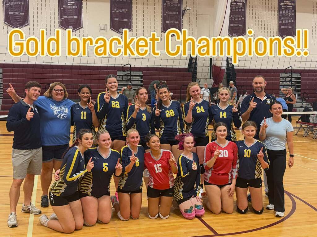 A team, likely of female volleyball players, poses for a photo on a court, with some kneeling and others standing, against a gymnasium background.