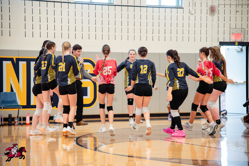 A group of female athletes in matching uniforms gather in a huddle on a gym floor.