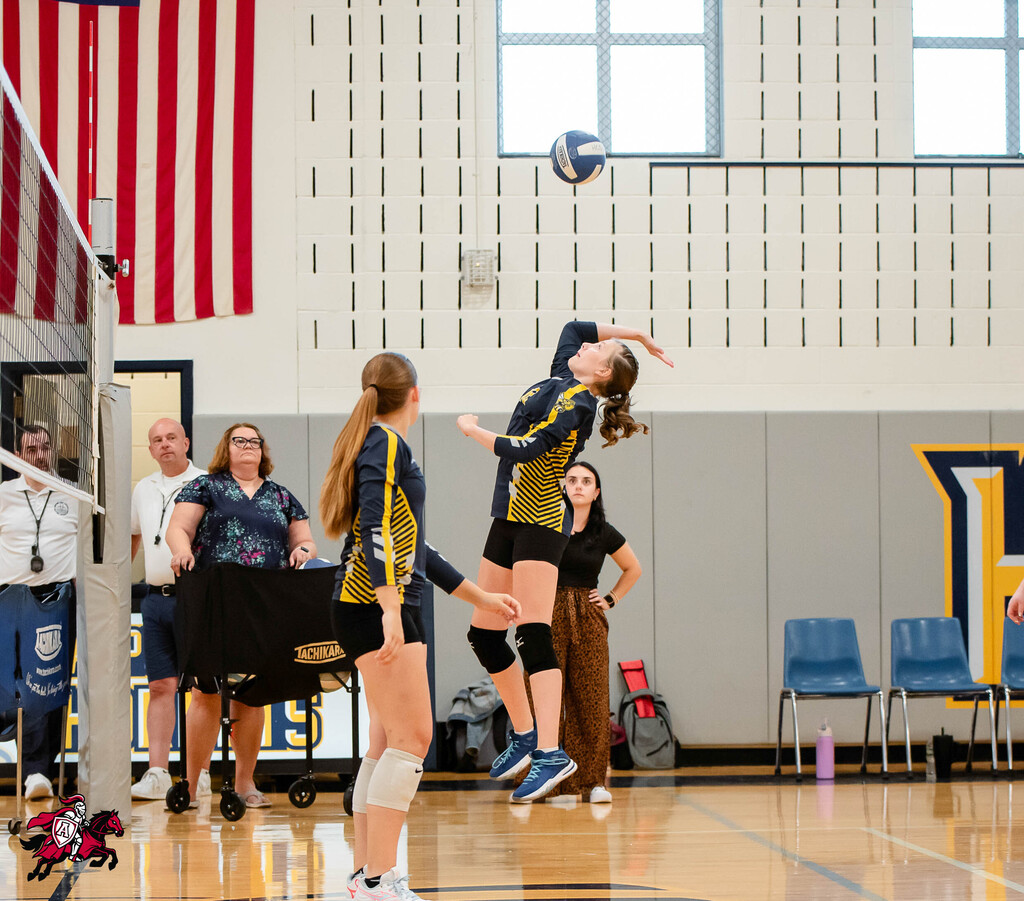 Two volleyball players, one jumping to hit a ball, others watching. Background includes an American flag, bleachers, chairs, and a yellow letter "L".