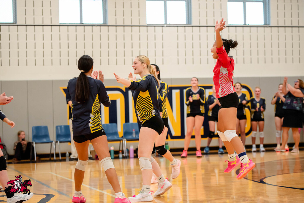 A group of female athletes in sportswear and knee pads playing volleyball indoors, one jumping to spike.