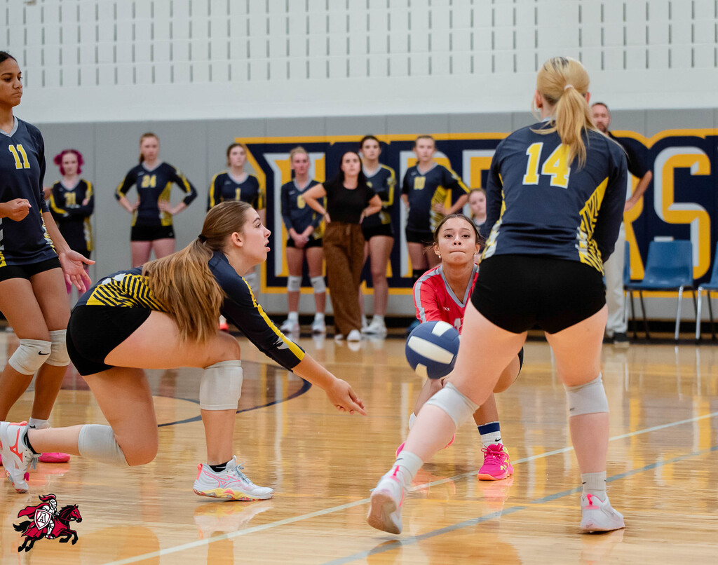 Volleyball players compete on a court, one woman dives to block a ball while another runs forward.