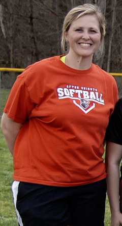 Lacy Hart wearing an orange t-shirt with "Softball" and a logo on it, smiling, with a grassy background and trees.