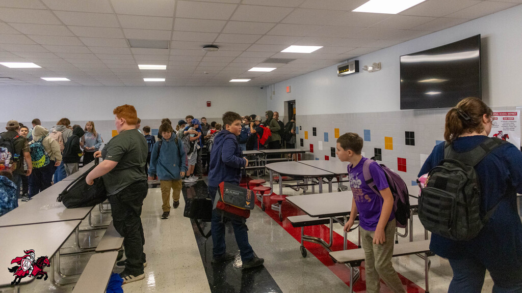 A room with students standing at desks, moving around and carrying bags. Some people have backpacks.