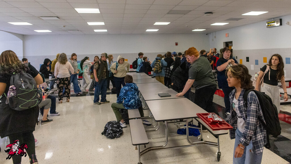 A room with tables and chairs where students are gathered, some standing, others seated. Backpacks and trays are visible.
