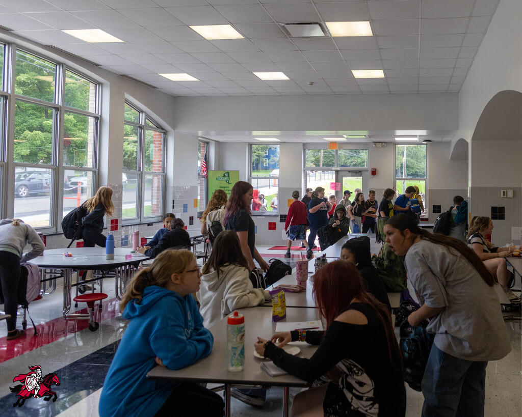 A cafeteria with multiple people seated and standing around tables. Bright sunlight filters through large windows.