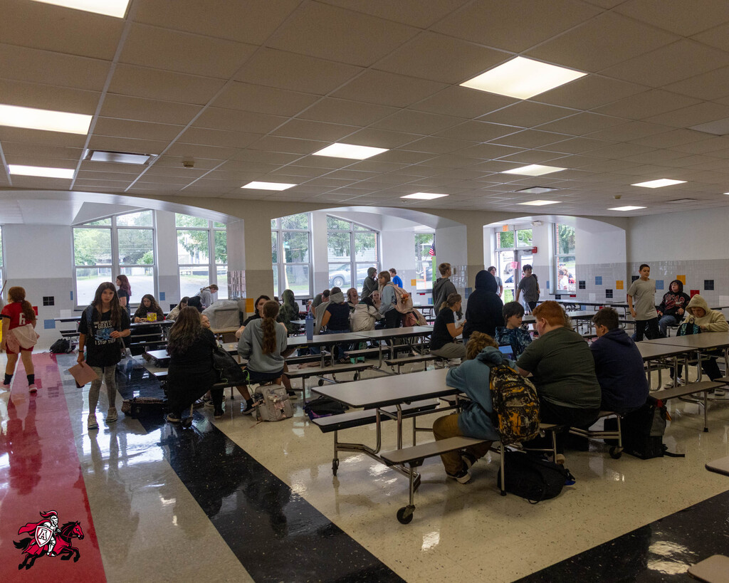 A large cafeteria with many tables and chairs. People are seated and standing. The floor has a red line.