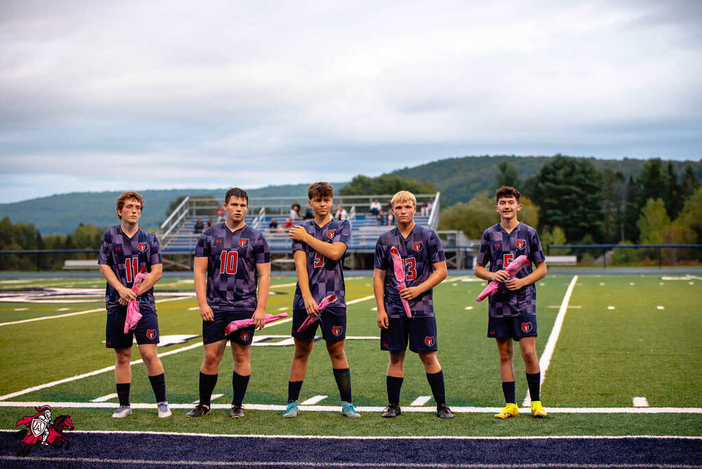 Five male soccer players stand on a field. They wear matching uniforms and hold pink items. A stadium and mountains are in the background.