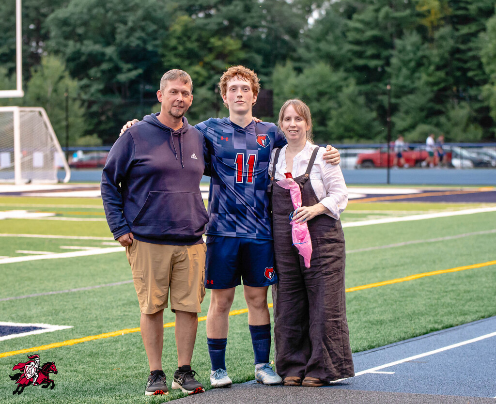 Three people stand on a soccer field with trees in the background. One wears a blue jersey, another a brown dress.