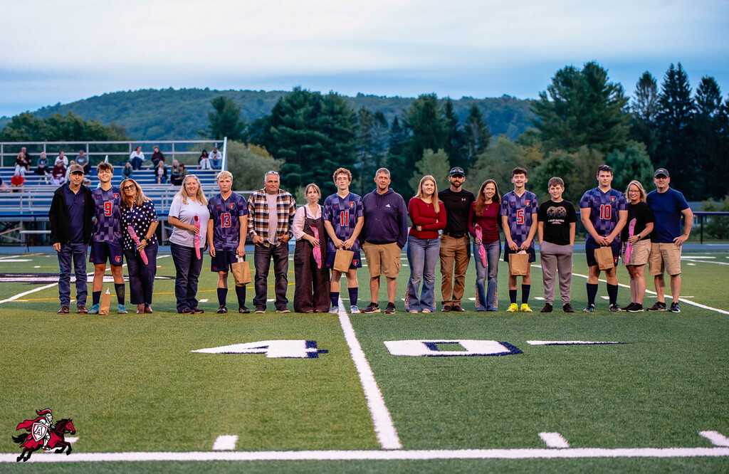 A group of adults and children stand on a field, wearing similar shirts and holding baseball bats.