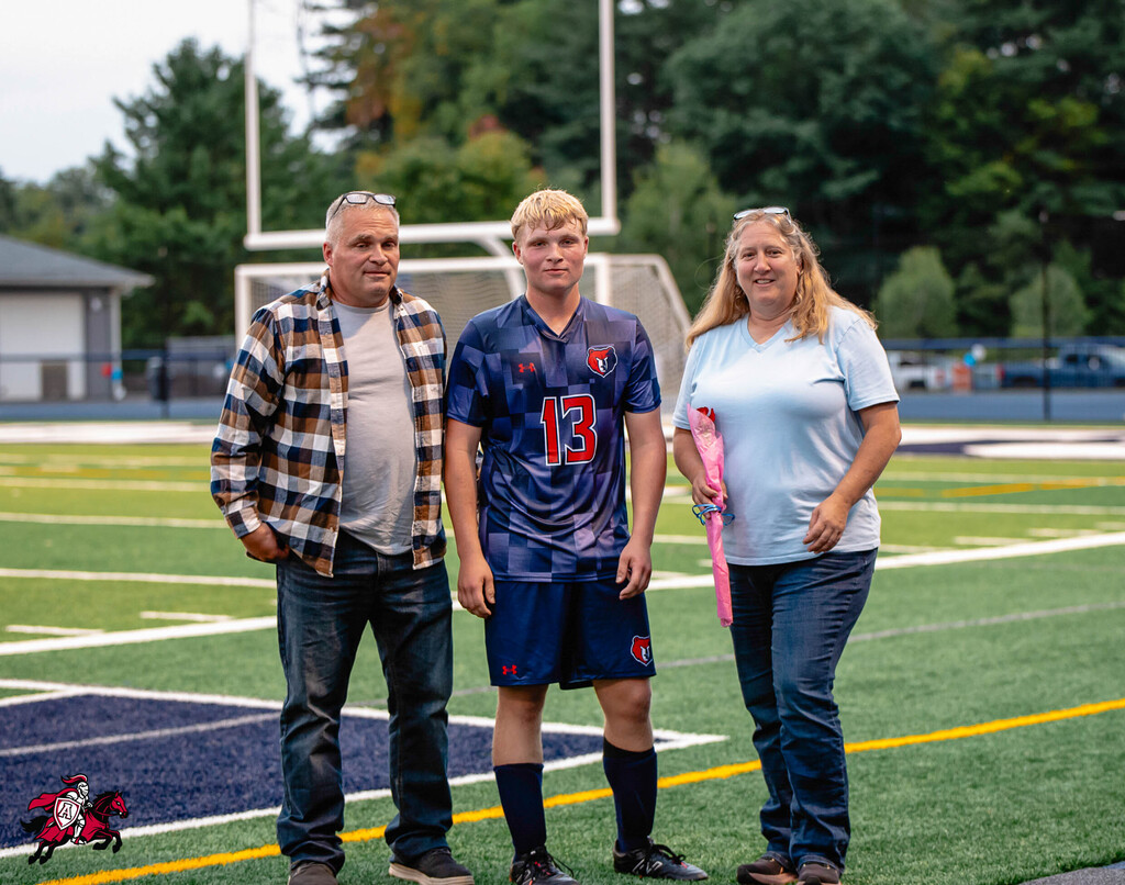 Three people on a football field: a young player in a blue uniform, a man in a plaid shirt, and a woman in a white shirt.