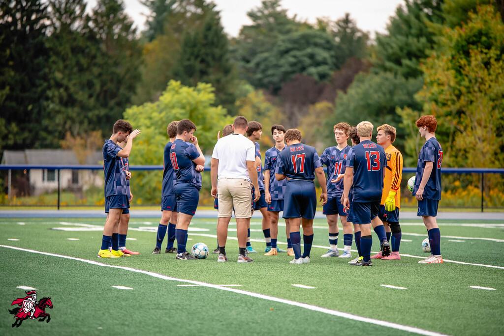 Players in blue uniforms gather on a soccer field, with a coach in white standing among them. Trees and a fence are in the background.