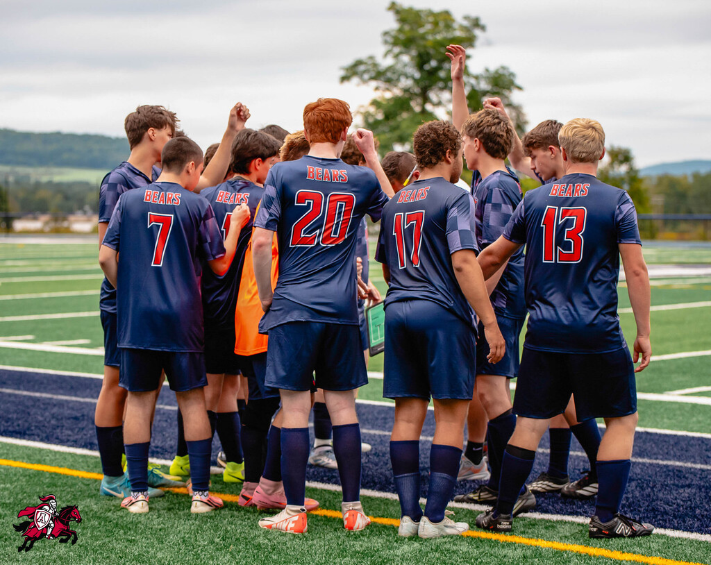 Team members in blue uniforms with numbers 7, 13, 17, 20, and 30 huddle on a field with mountains in the distance.