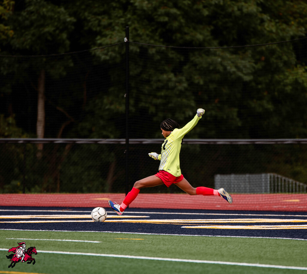 A soccer player kicks the ball on a field with a red and black border. Trees in the background.