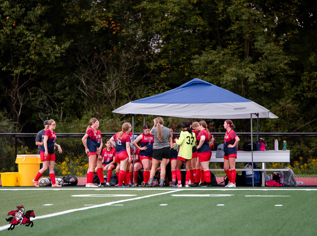 A soccer team in red uniforms gathers under a blue tent on a field, with trees and a fence in the background.
