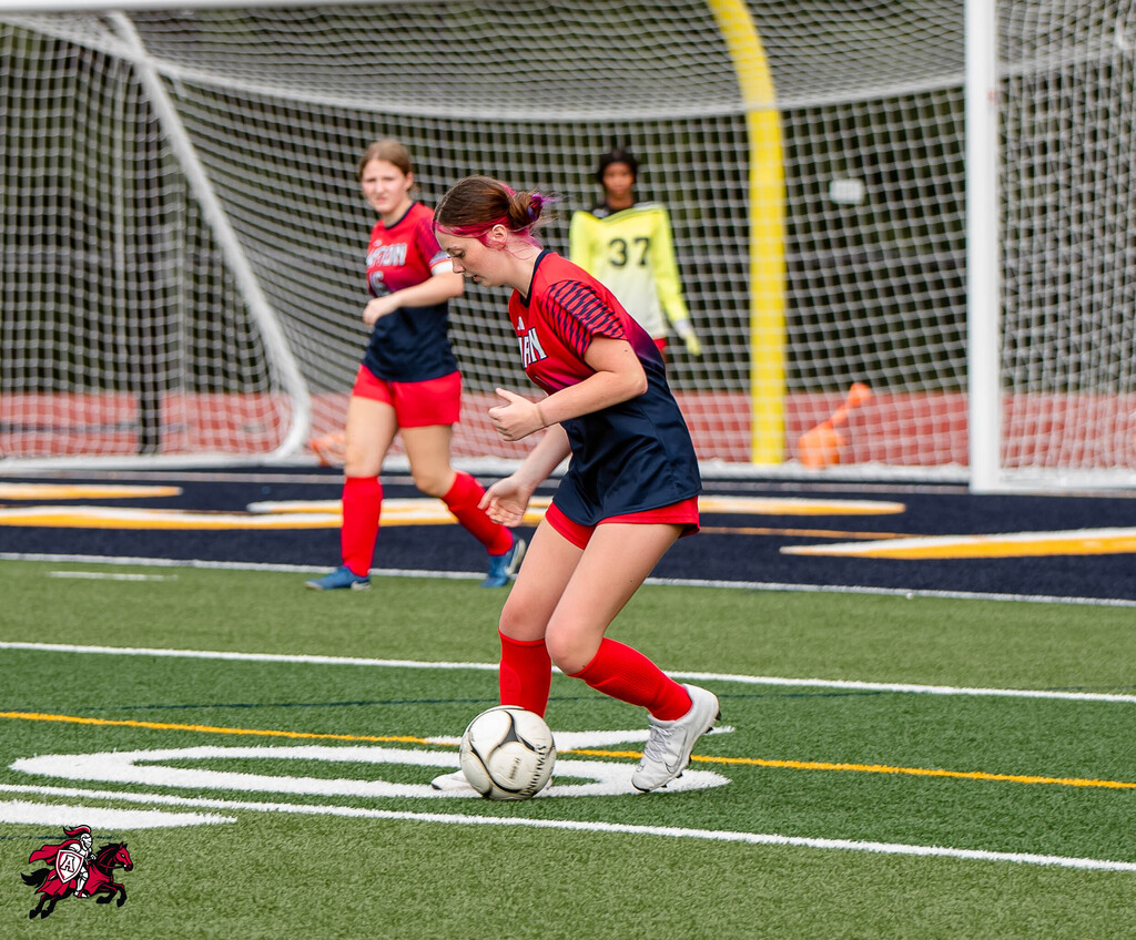Three female soccer players on a field. One player in red and blue is kicking a ball.