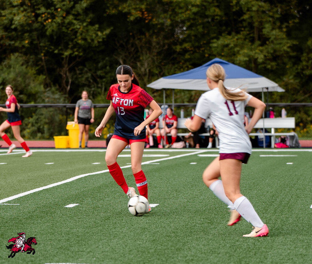 Two women playing soccer. One in red and black, the other in white and maroon. Trees in background.