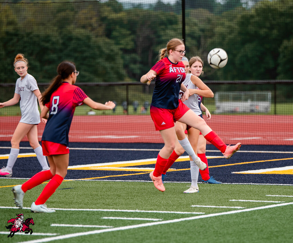Four female athletes in team jerseys play soccer on a field with white lines, a fence, and trees in the background.
