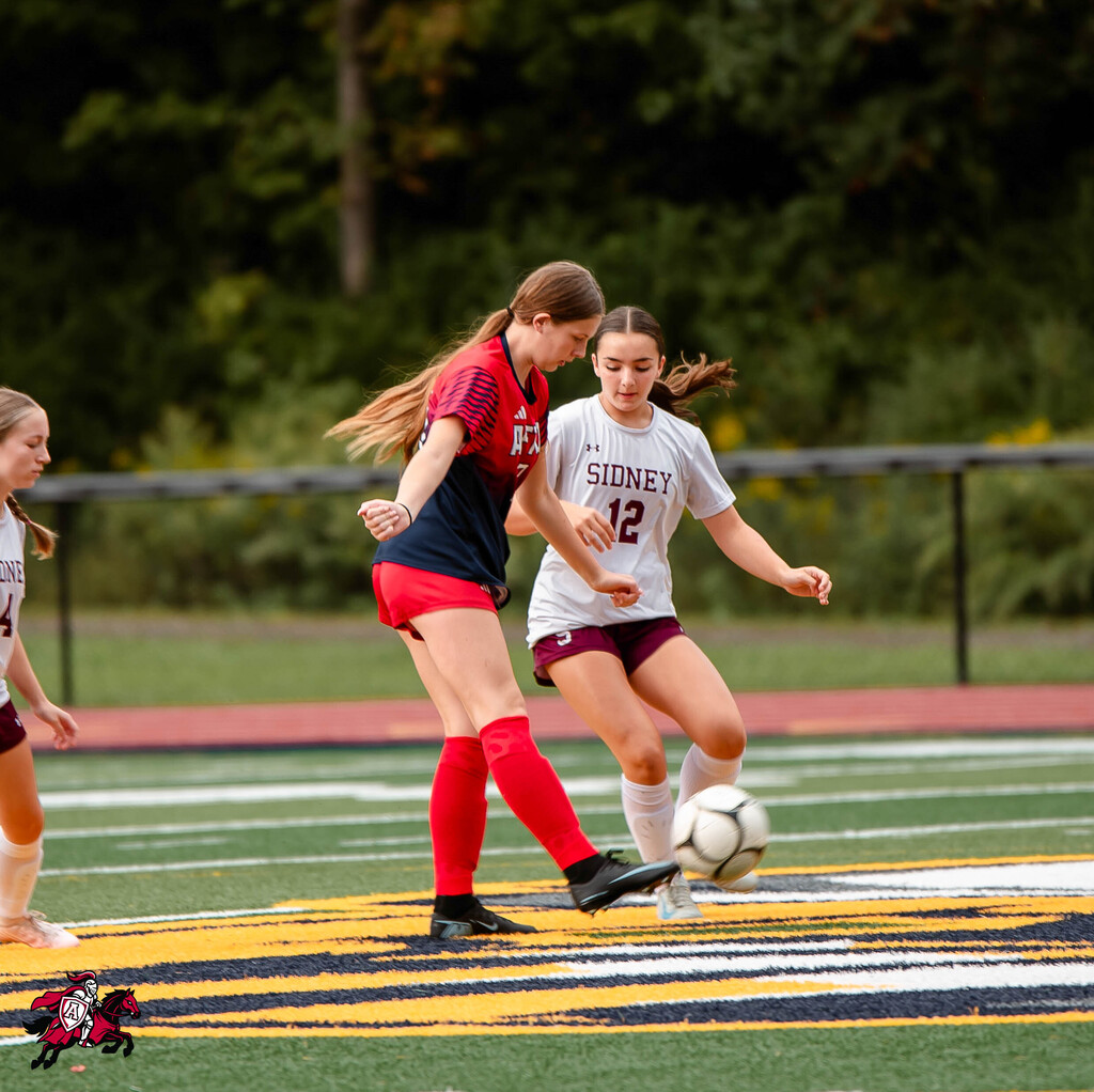 Three players in uniforms on a field, one with "SIDNEY" and number 12, playing soccer.