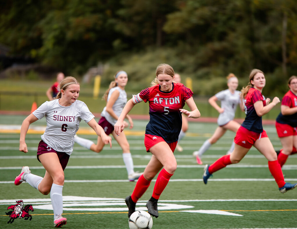 Soccer players in maroon and navy uniforms run on a field, chasing a white ball. Trees form the background.