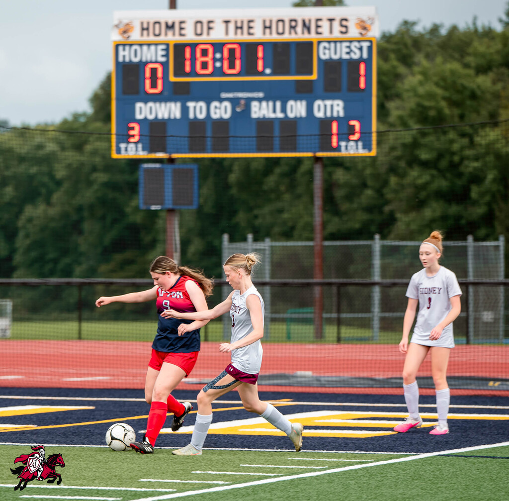 Three players on a soccer field; one chasing a ball, two looking on, with a scoreboard.