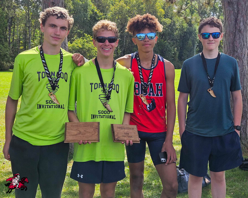 Four young men stand in a park, holding trophies and medals. Two wear green shirts; one has "Tough Mudder" on his.