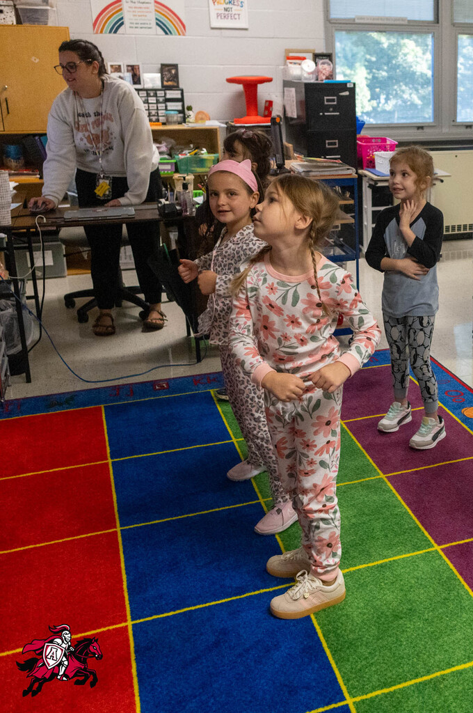 Four young girls in pajamas on a multicolored mat, one looking upward, a teacher in background.