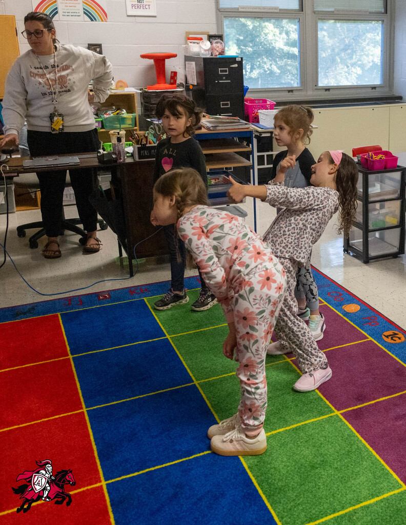 Five children play on a colorful floor mat while a teacher stands nearby. They wear pajamas.