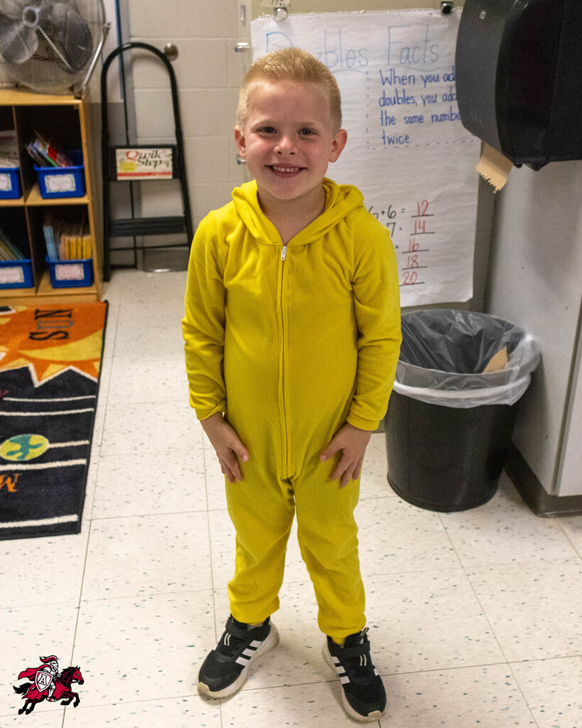 A young boy stands in a classroom, wearing a yellow jumpsuit, with a trash can, a whiteboard, and shelves in the background.