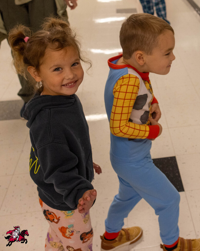 Two children, a girl in a blue hoodie and a boy in blue and yellow pajamas, smile. Behind them, two people walk in the hallway.