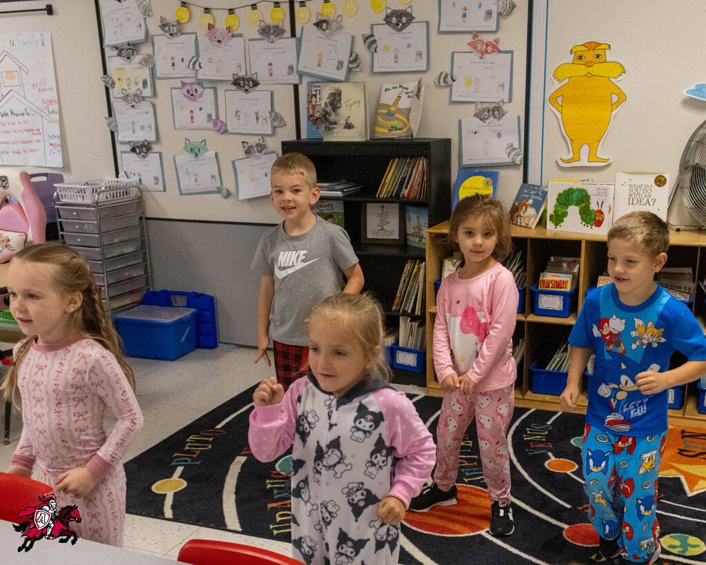 Kids in pajamas stand in a classroom with a rug, bookcase, and bulletin board. Four children face front.