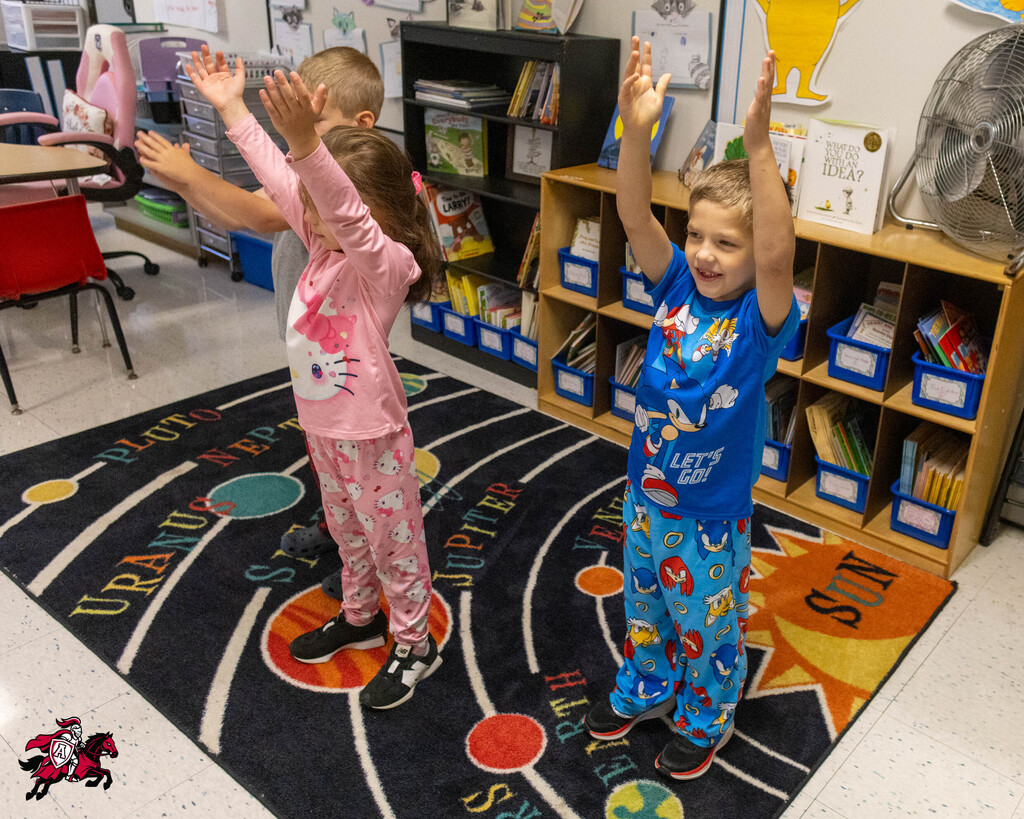 Children in pajamas raise their hands above their heads on a rug depicting the solar system.