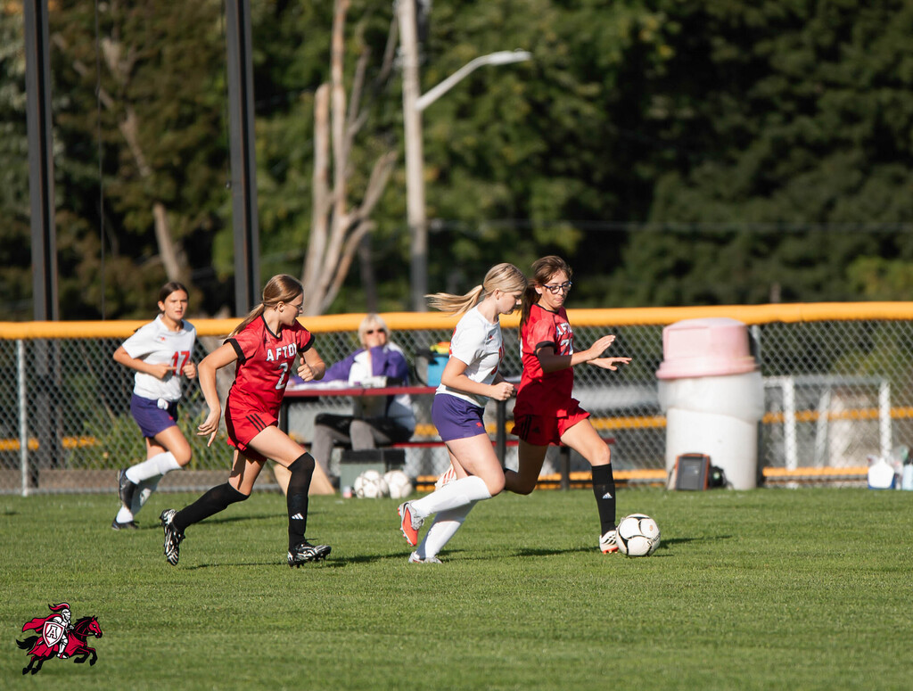 Four women play soccer on a field. Two wear red, one white, one purple. Spectators sit behind a fence.