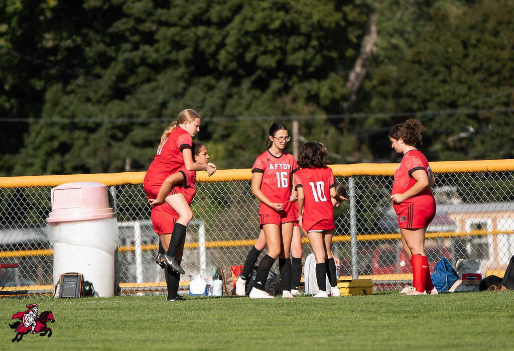 Soccer players in red uniforms on a field, with one jumping in the air, others standing, and a fence behind them.