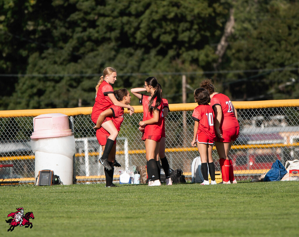 Women's soccer team on a field, with one player lifted by others. Behind them, a fence and a trash can.