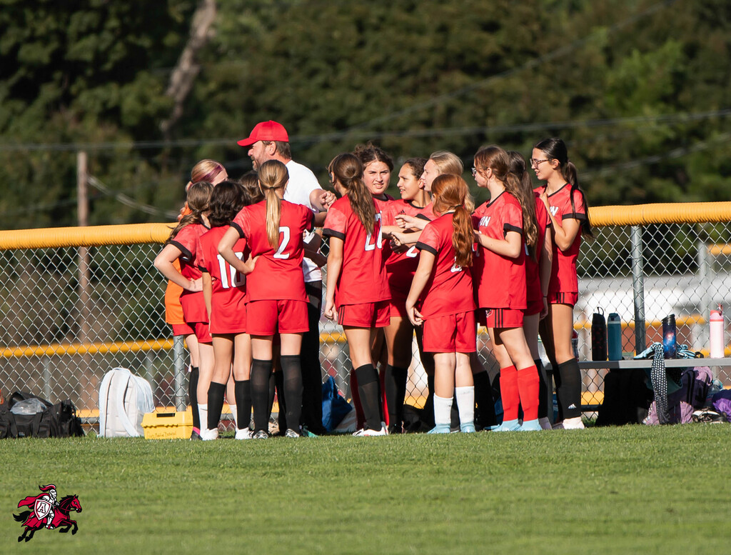 Soccer players in red uniforms gather for a team talk near a yellow fence with a grassy field.