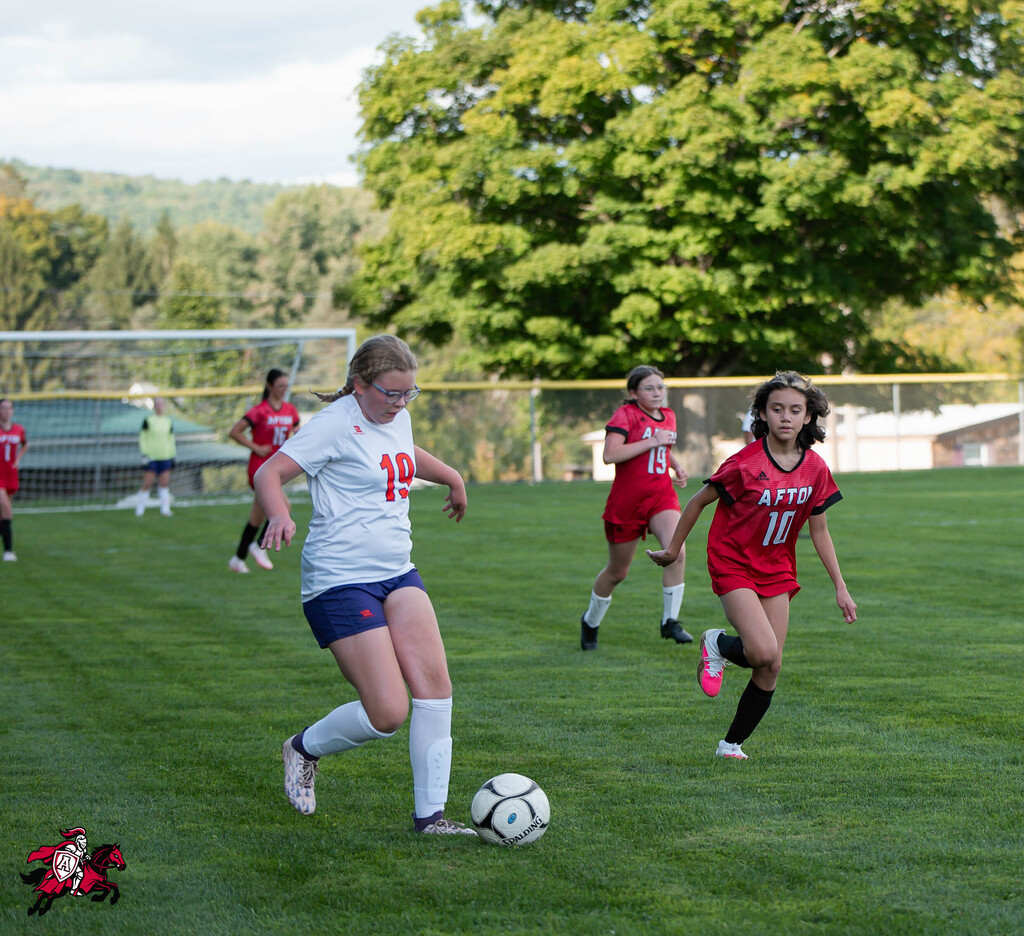 On a soccer field, one player runs with the ball. Other players in red and white uniforms are in the background.
