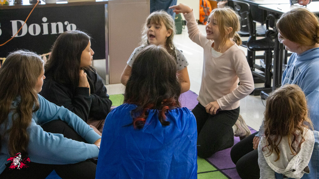 A group of children and adults in a classroom. A young girl raises her hand and talks.