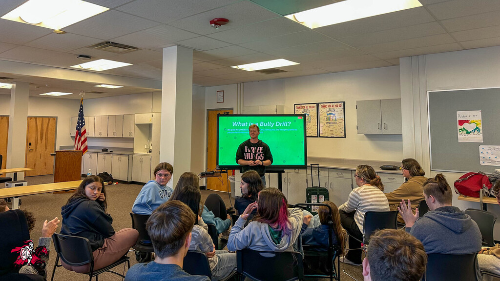 A classroom with a large screen displaying a man. Attendees sit in chairs, facing the screen.