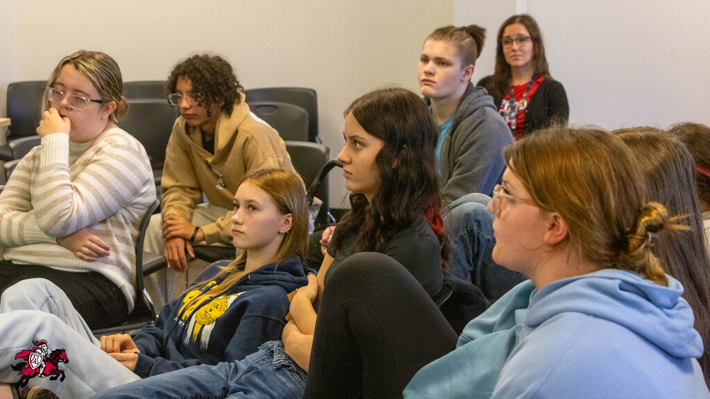 Nine individuals sit in chairs, facing forward, in a room. Two wear glasses. Some have their arms crossed.