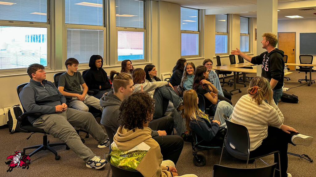A group of people sit around a circle, engaged in a discussion with a person standing in the center.