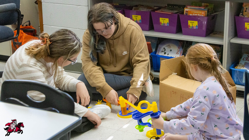 Three people work with toys on a floor. A woman sits in the middle, and two children are on her sides.