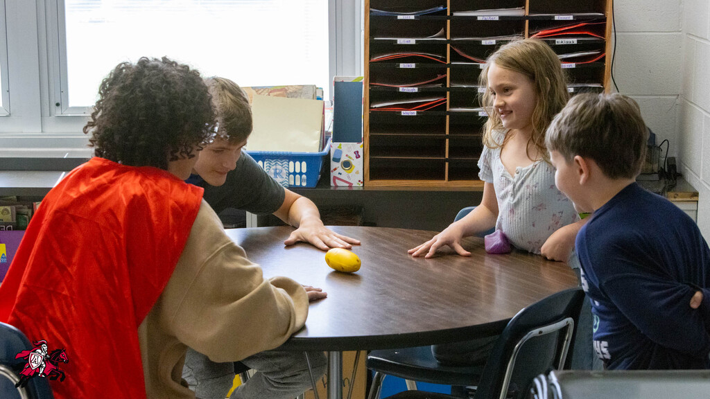 Four children sit around a table. Two girls, one wearing red, hold an orange ball. A boy leans forward.