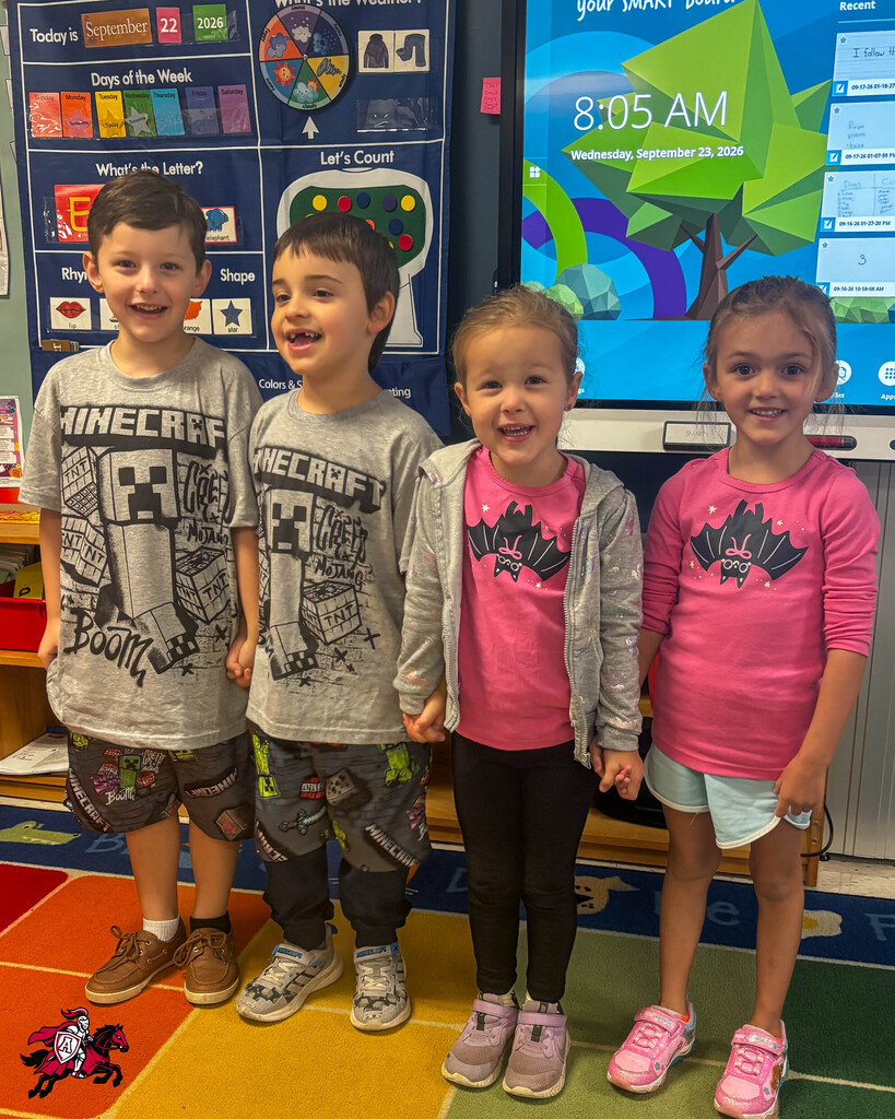Four children in a classroom, smiling, standing in front of a monitor displaying a green screen.