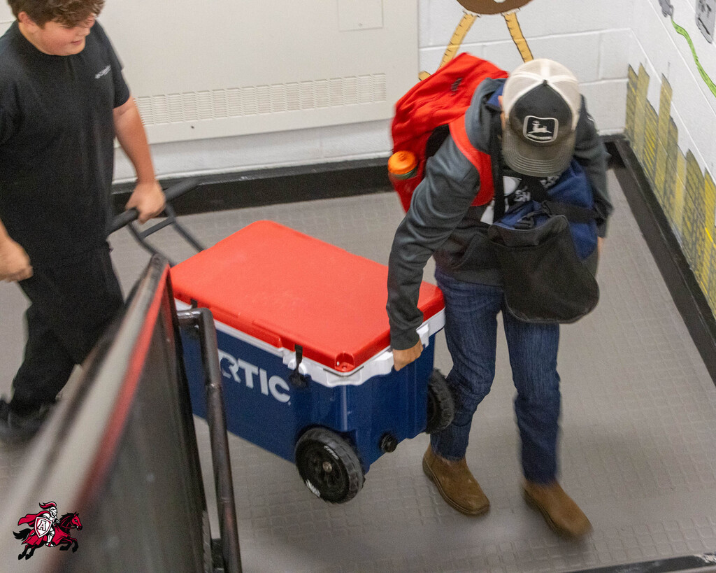 Two people pushing a cooler cart up an escalator. One wears black, the other wears blue jeans and a cap.